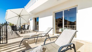 A sunlit outdoor seating area with lounge chairs and a table, a white umbrella, and a glass door leading to a covered patio.