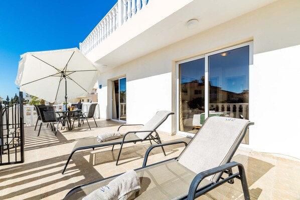 A sunlit outdoor seating area with lounge chairs and a table, a white umbrella, and a glass door leading to a covered patio.