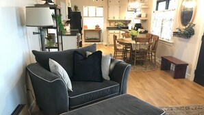 A living room with a grey sofa and ottoman, a dining table with chairs, and a kitchen in the background.
