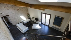 Een moderne loft-achtige kamer met een stenen muur, een witte bank, een klein tafeltje en een televisie. Er is een trap die naar een hoger niveau leidt.