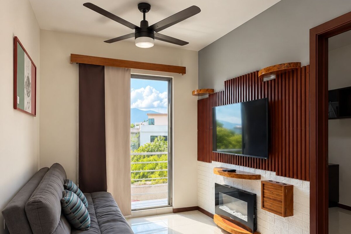 Una sala de estar moderna con un sofá, un televisor de pantalla plana montado en un panel de madera, un ventilador de techo y un ventanal con vistas a edificios.