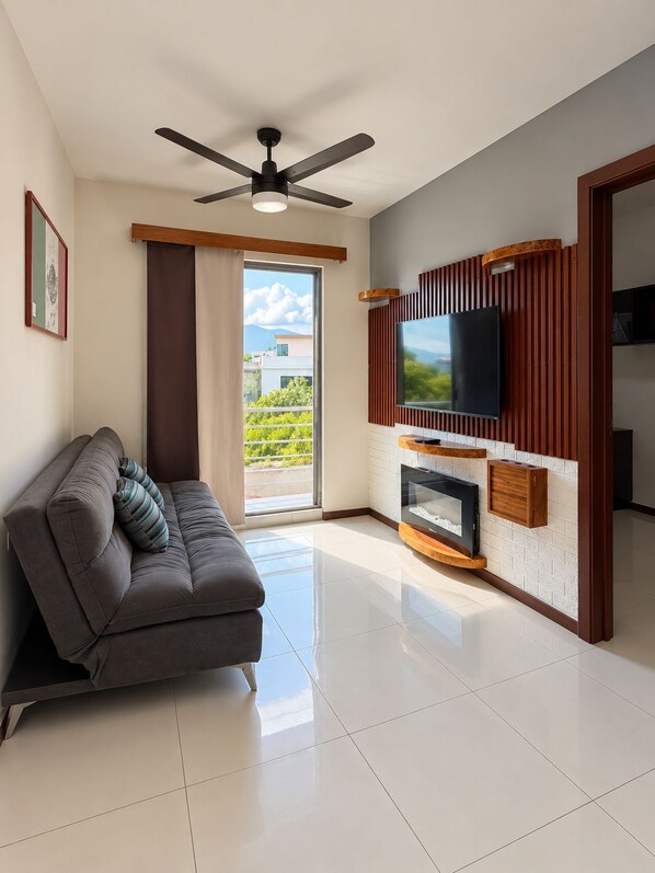 Una sala de estar moderna con un sofá, un televisor de pantalla plana montado en un panel de madera, un ventilador de techo y un ventanal con vistas a edificios.