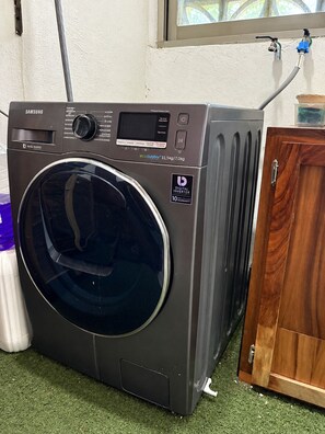 Una lavadora Samsung con tecnología digital Inverter y garantía de 10 años, ubicada sobre un piso de césped artificial verde junto a un armario de madera.