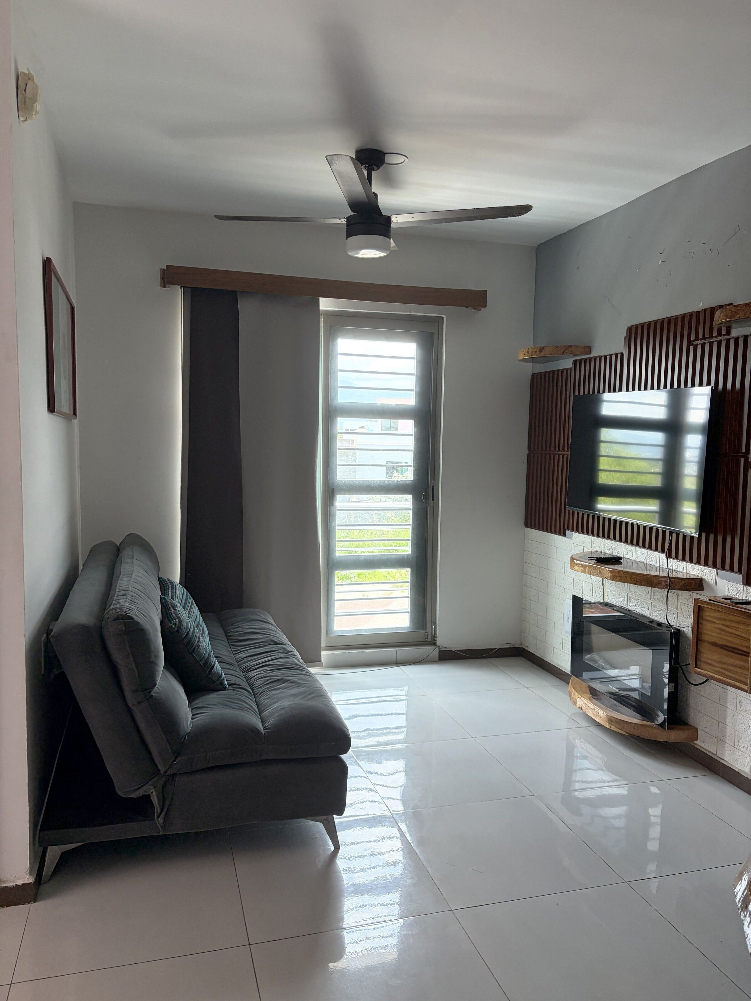 Una sala de estar moderna con un sofá gris, un ventilador de techo, un ventanal grande y un televisor de pantalla plana montado en una pared de madera.