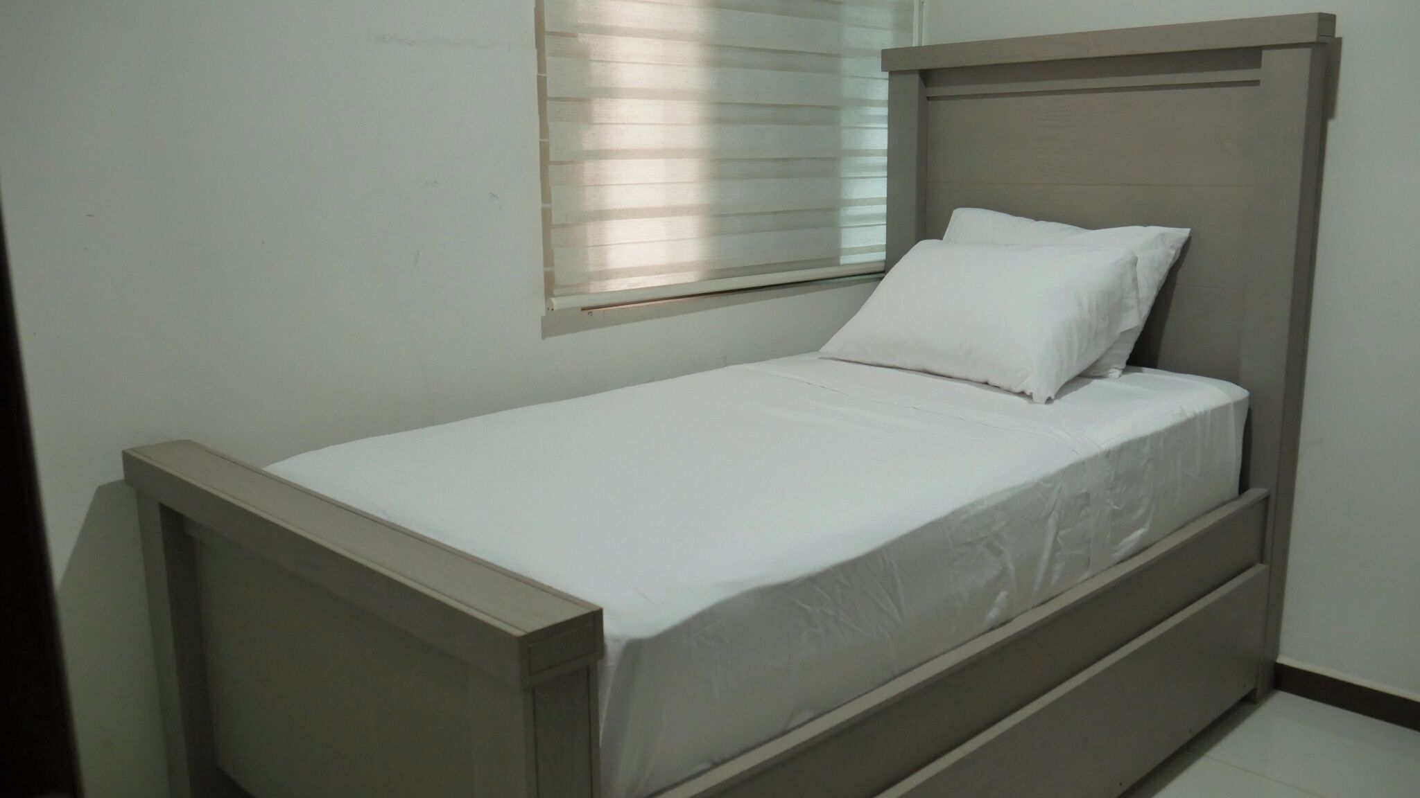A single bed with a wooden frame and white bedding in a room with a window with blinds.