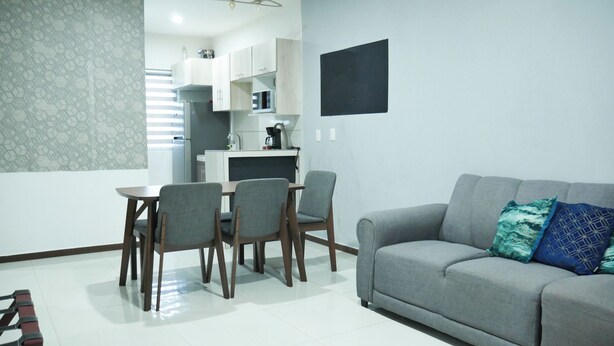 Ein modernes Wohnzimmer mit einem grauen Sofa, einem Esstisch mit vier Stühlen und einer Küchenzeile mit weißen Schränken und einem schwarzen Wand-Fernseher.