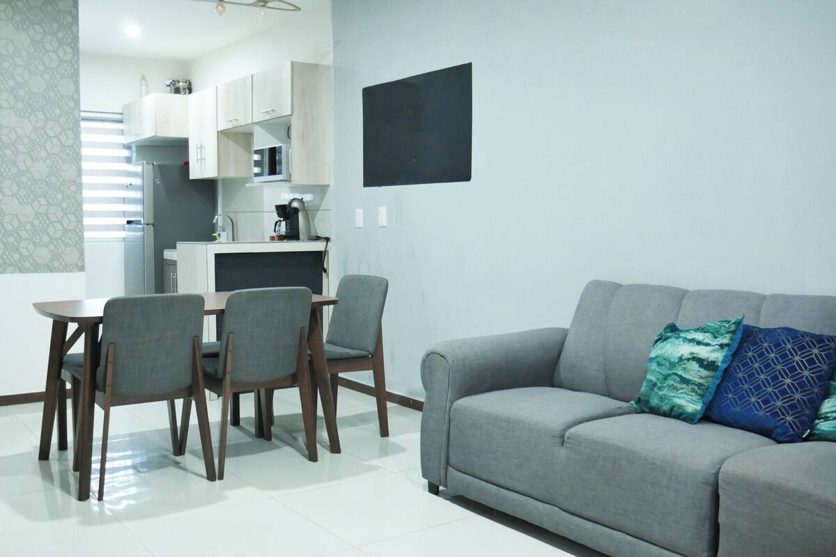 Un salon moderne avec un canapé gris, une table à manger avec quatre chaises et un coin cuisine doté de meubles blancs et d’un téléviseur noir fixé au mur.