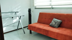 Una habitación con un escritorio blanco, un ventilador de pie negro y un sofá naranja con un cojín.