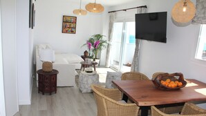 Un salon moderne avec un canapé, une table à manger entourée de chaises en osier, un téléviseur fixé au mur et une grande fenêtre offrant une vue sur la mer.