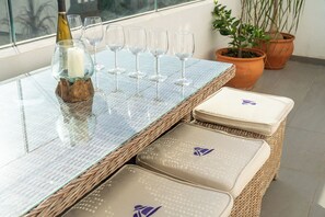 Ein Tisch mit Glasplatte, Weingläsern und einer Kerze, Korbo ttomanen mit Kissen sowie Topfpflanzen.
