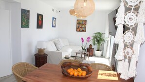 Un coin repas avec une table en bois, des chaises en osier et un saladier rempli d’oranges. On aperçoit un canapé blanc et une plante en pot à l’arrière-plan.
