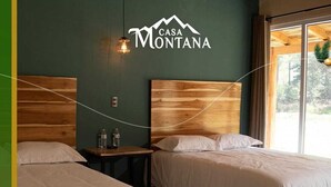 A hotel room with two beds, wooden headboards, a wall-mounted sign reading "CASA MONTANA", and a window with a view of greenery.