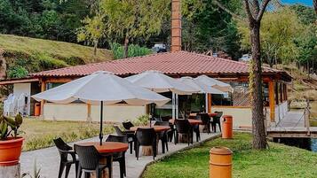 An outdoor dining area with tables and chairs, umbrellas, and a building with a red roof in the background.