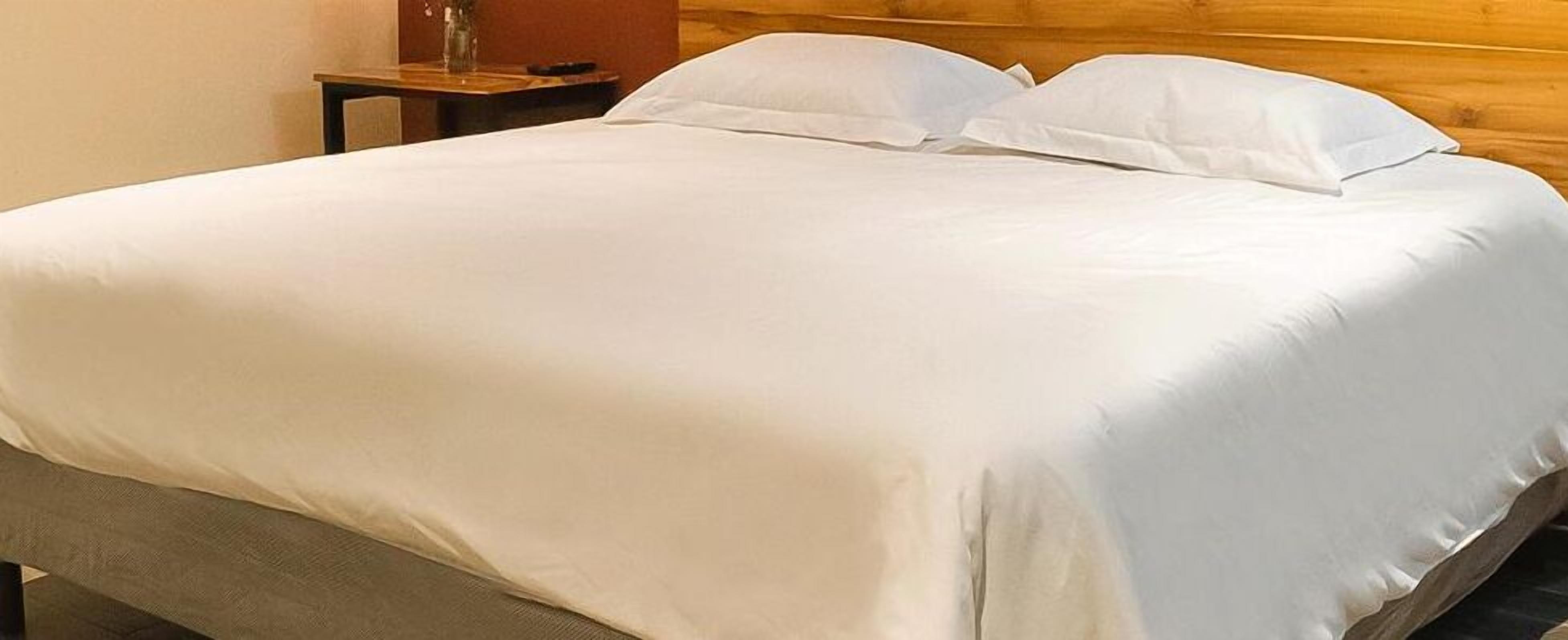 Un lit bien fait, avec du linge de lit blanc, dans une chambre dotée d’une tête de lit en bois et d’une petite table sur laquelle est posé un vase.