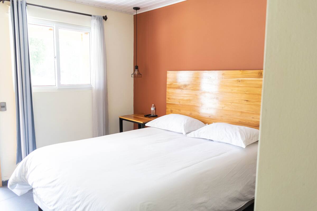 Una cama bien hecha con sábanas blancas en una habitación con cabecera de madera y una mesita de noche pequeña.