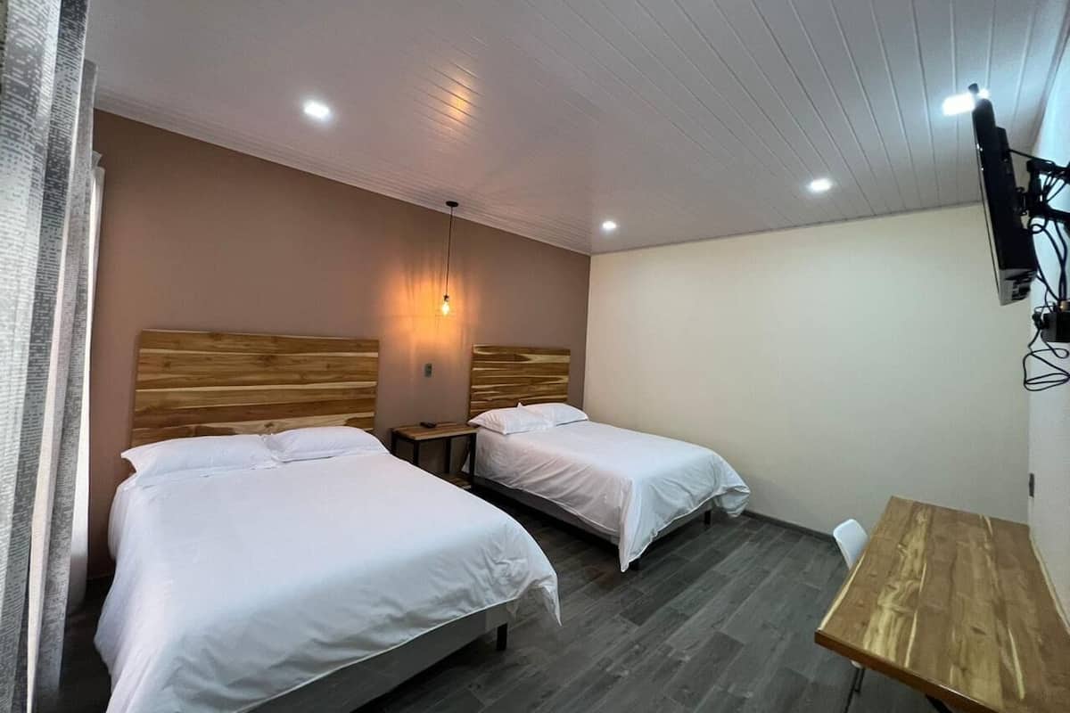 Habitación de hotel con dos camas, cabecero de madera, una mesita pequeña y televisor montado en la pared.
