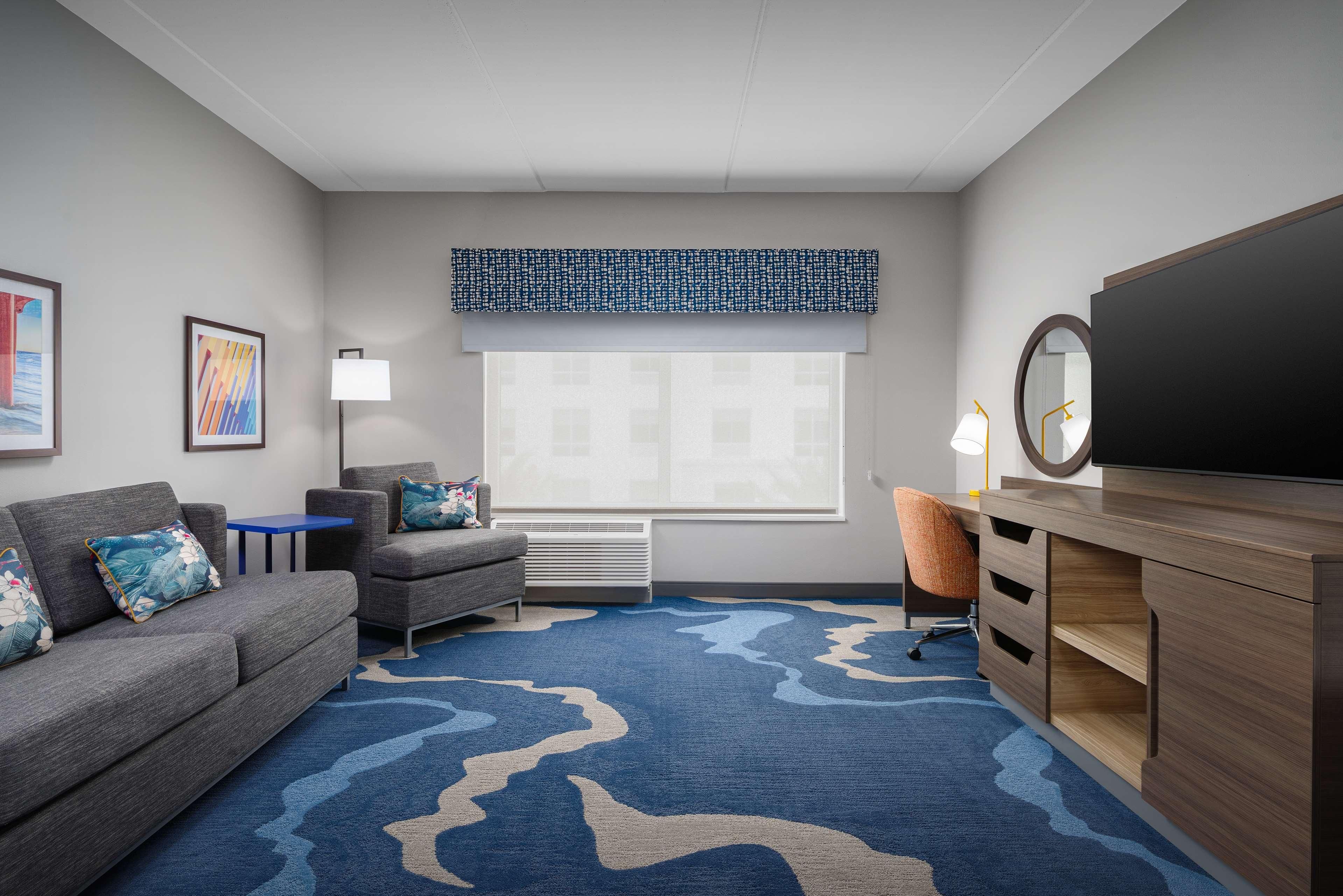 A modern hotel room with a grey sofa, a blue patterned rug, a wooden desk, and a large window with blinds.
