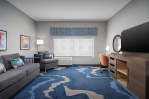 A modern hotel room with a grey sofa, a blue patterned rug, a wooden desk, and a large window with blinds. - Hampton Inn And Suites By Hilton Naples South (Naples)