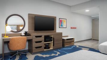 Habitación de hotel con una pantalla plana grande, un escritorio de madera, una silla, un microondas, una cafetera y una alfombra azul estampada.