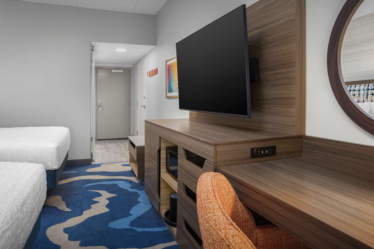 Ett hotellrum med en stor platt-TV som hänger på väggen, ett skrivbord i trä med lådor, en bekväm orange fåtölj och två sängar med vita sängkläder.
