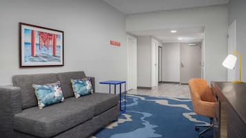 Una habitación de hotel moderna con un sofá gris, cojines estampados con motivos florales, una alfombra azul, un escritorio de madera y una silla naranja.