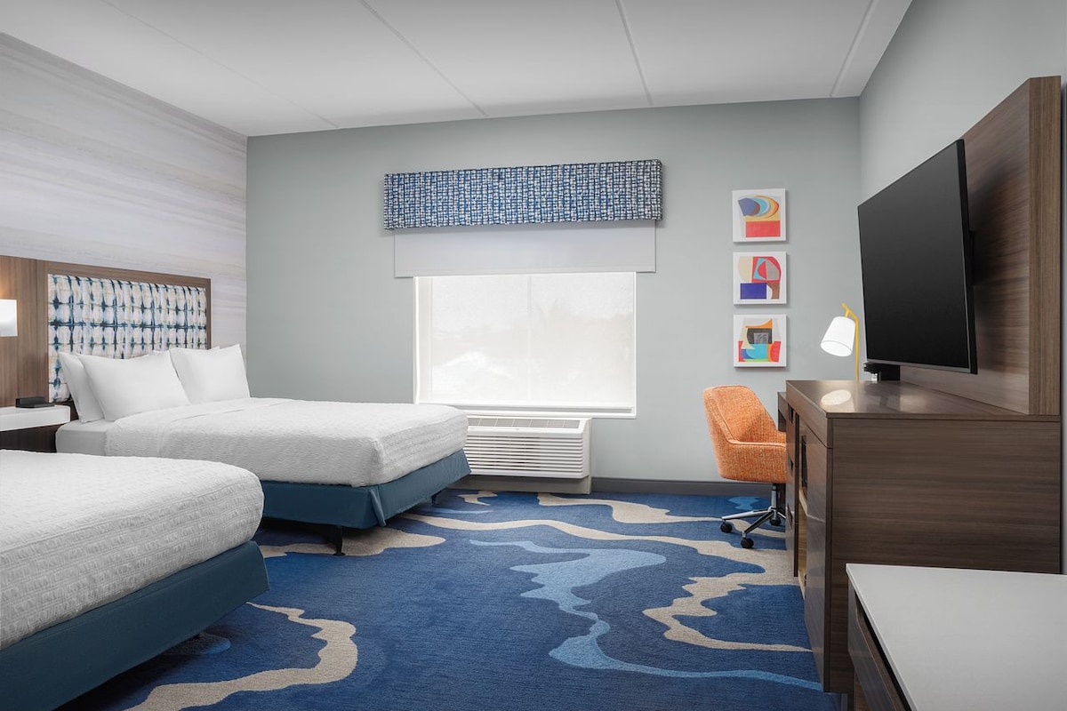 A hotel room with two beds, a large window, a flat-screen TV, a desk, and a chair.