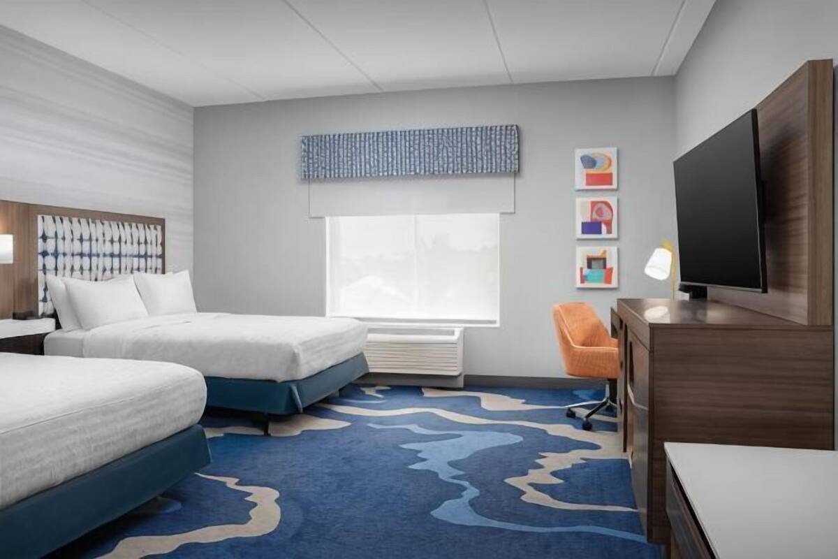A hotel room with two beds, a flat-screen TV, a desk, a chair, and a large window.