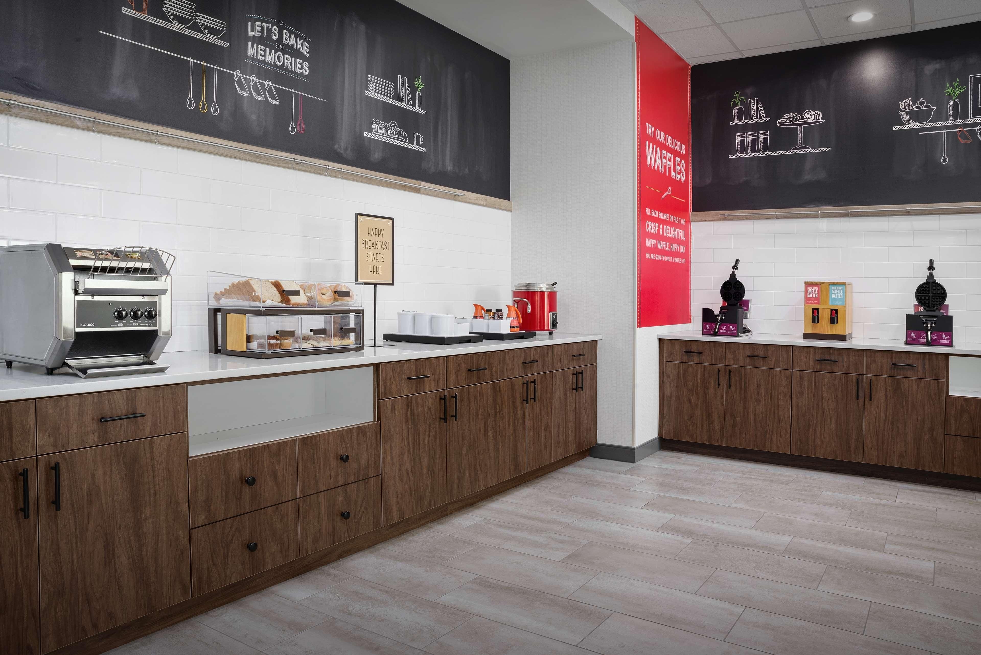 Una cocina moderna con armarios de madera, una cafetera, máquina para gofres y una pared de pizarra con dibujos relacionados con el desayuno.
