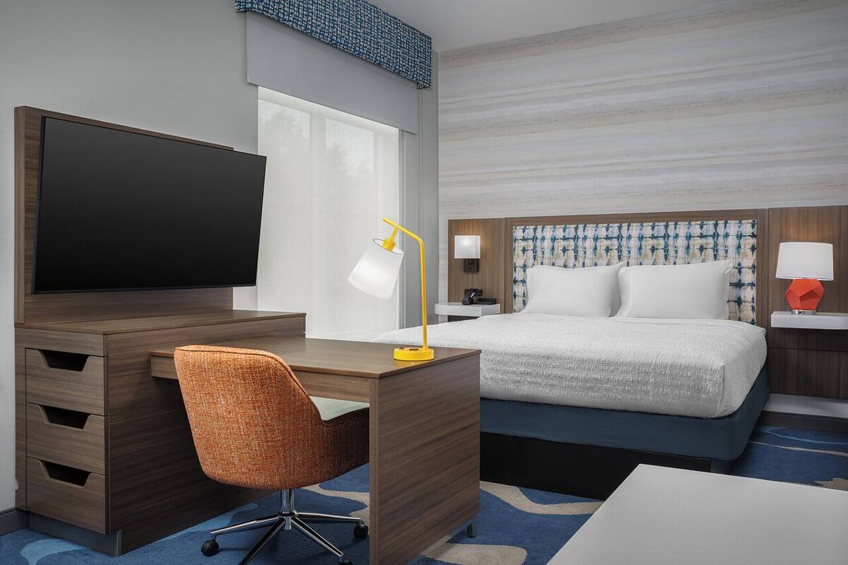 A hotel room with a wooden desk, a television, a chair, a bed with white bedding, and a nightstand with a lamp.