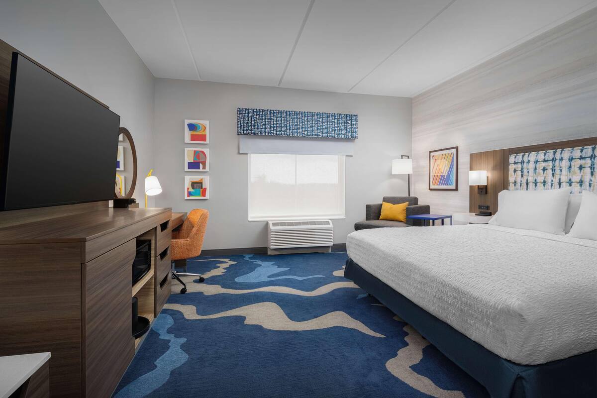 A hotel room with a bed, a desk, a chair, a TV, a window with blinds, and artwork on the walls.