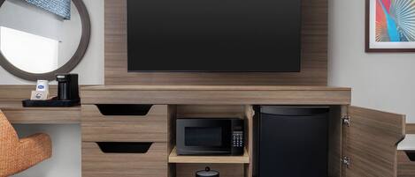 Une chambre d’hôtel moderne équipée d’un téléviseur à écran plat fixé au mur, d’un four à micro-ondes, d’un réfrigérateur et d’une machine à café, le tout posé sur une console en bois.