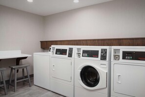 A laundry room with three stacked front-loading washing machines, a wooden countertop, and two stools. - Hampton Inn And Suites By Hilton Naples South (Naples)