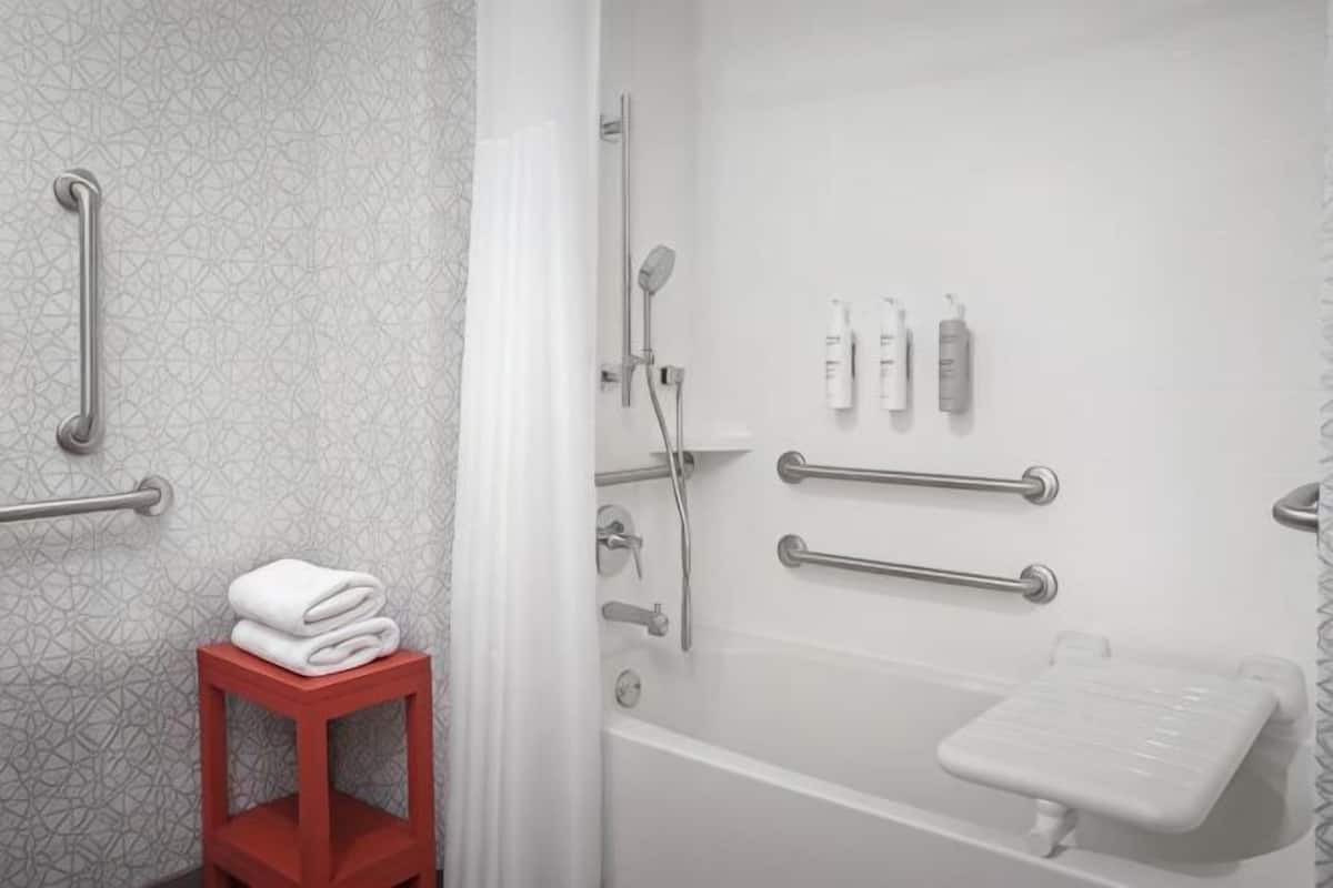 A bathroom with a shower, grab bars, a red stool, and a white sink.