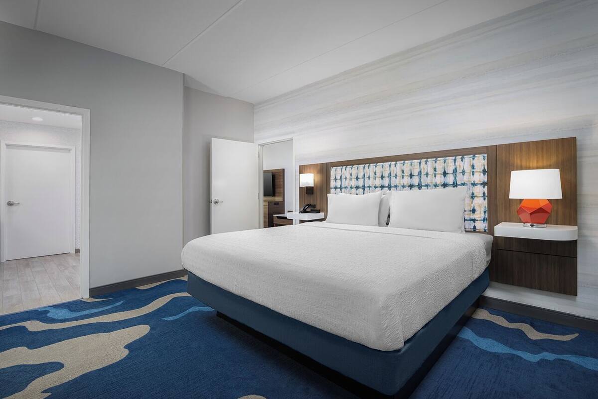 A hotel room with a large bed, a bedside table, a nightstand, a mirror, and a wall-mounted lamp.