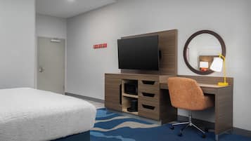 A hotel room with a bed, a desk with a chair, a television, a mirror, and a small lamp.