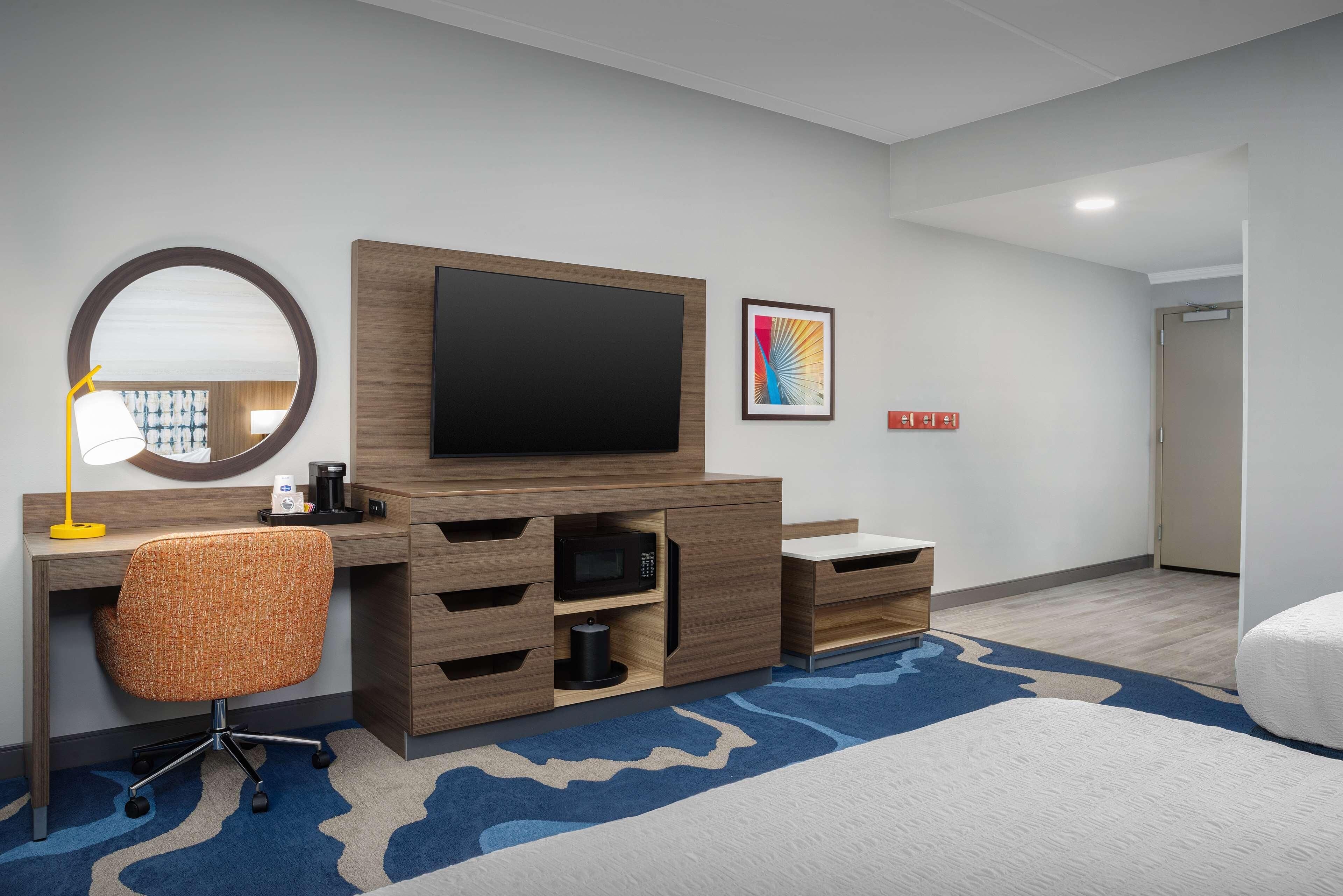 Hotelkamer met een houten bureau, een flatscreen televisie, een ronde spiegel, een magnetron, en een bed met een vloerkleed met blauw-witte patronen.