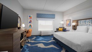 Habitación de hotel con cama, escritorio, silla, televisor, ventana con persianas y obras de arte en las paredes.