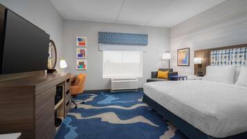 Ein Hotelzimmer mit Bett, Schreibtisch, Stuhl, Fernseher und einem Fenster mit Jalousien. Kunstwerke sind an den Wänden angebracht.