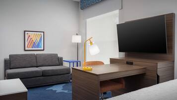 A modern hotel room with a grey sofa, a wooden desk, a flat-screen TV, a lamp, and a framed abstract art piece on the wall.