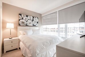 Une chambre d’hôtel avec un grand lit, une table de chevet avec une lampe, un tableau au mur et une fenêtre avec des stores.