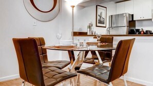 Un comedor moderno con una mesa de vidrio y sillas de cuero, una cocina con gabinetes blancos y electrodomésticos de acero inoxidable, y un espejo redondo en la pared.