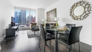 A modern living room with a glass dining table, black chairs, and a city view through large windows.