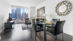 A modern living room with a glass dining table, black chairs, and a city view through large windows.