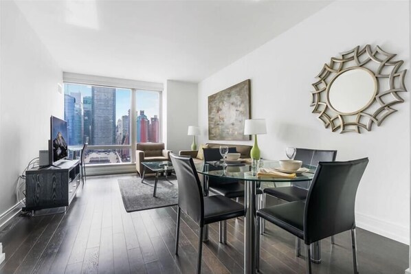 A modern living room with a glass dining table, black chairs, and a city view through large windows.