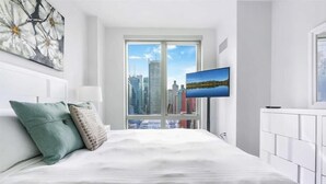 Una habitación de hotel moderna con una cama grande, vistas a la ciudad a través de un ventanal amplio y un televisor de pantalla plana montado en la pared.
