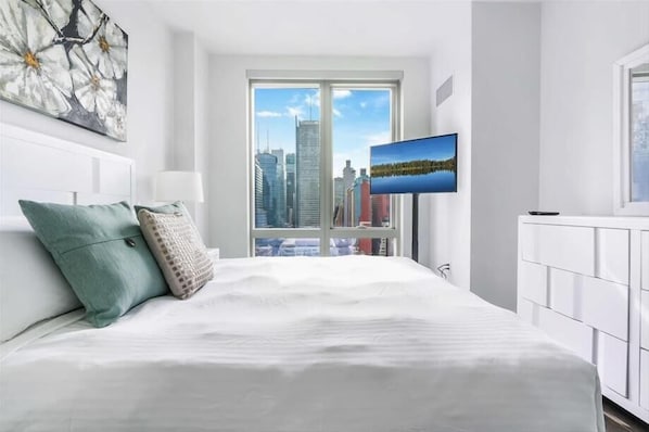 A modern hotel room with a large bed, a city view through a large window, and a flat-screen TV mounted on the wall.