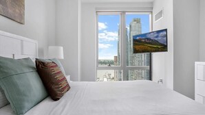 Una habitación de hotel moderna con una cama grande, un televisor de pantalla plana montado en la pared y vistas a la ciudad a través de amplias ventanas.