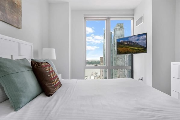 Una habitación de hotel moderna con una cama grande, un televisor de pantalla plana montado en la pared y vistas a la ciudad a través de amplias ventanas.