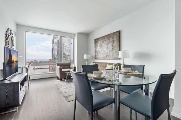 A modern living room with a glass dining table, comfortable seating, a large window with a city view, and a flat-screen TV. - Hell’s Kitchen Condo w/ Skyline Views (New York)
