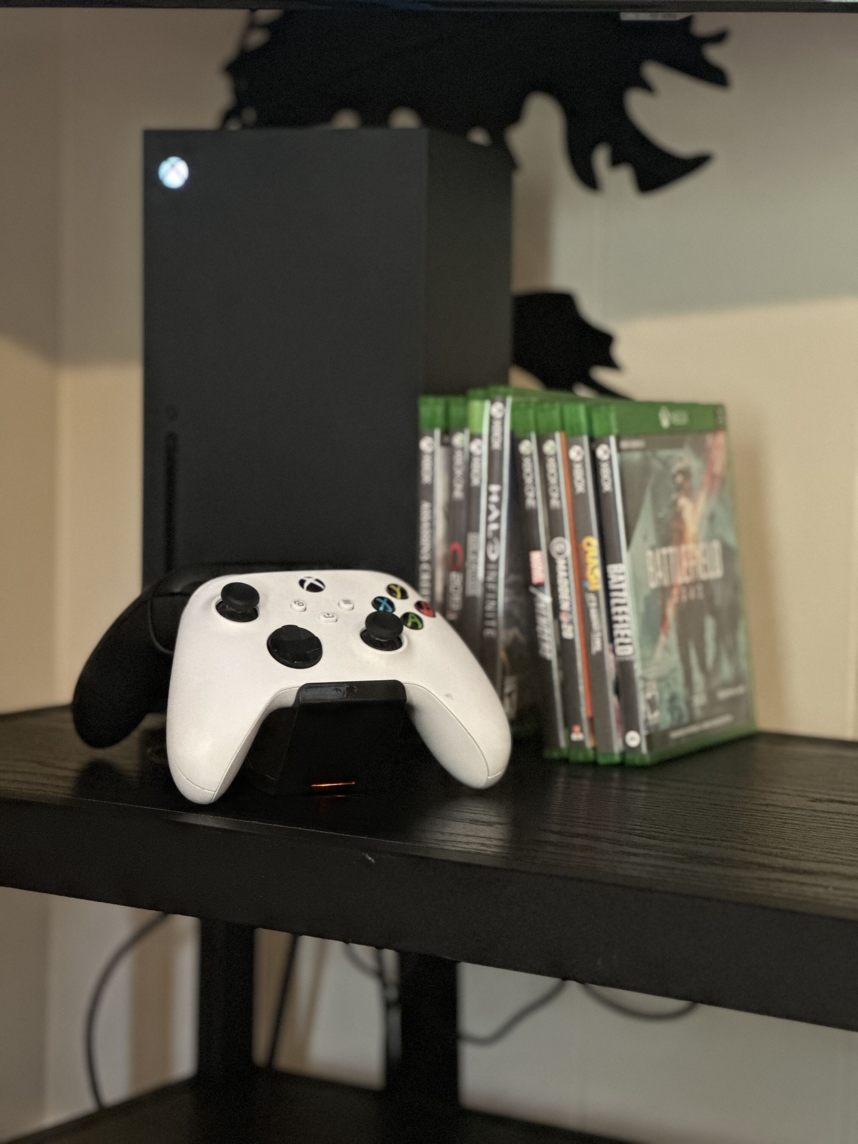 Un ensemble de jeu comprenant une manette Xbox blanche, une console de jeu noire et une rangée de boîtiers de jeux vidéo verts sur une étagère noire.